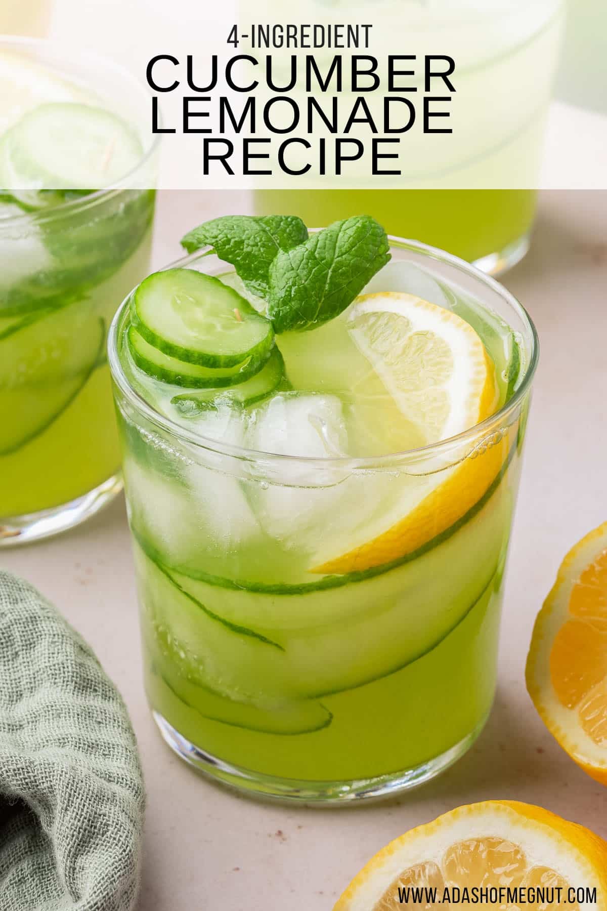 A glass of light green lemonade with cucumber ribbons, mint, ice cubes and a lemon wedge with additional glasses of lemonade in the background.
