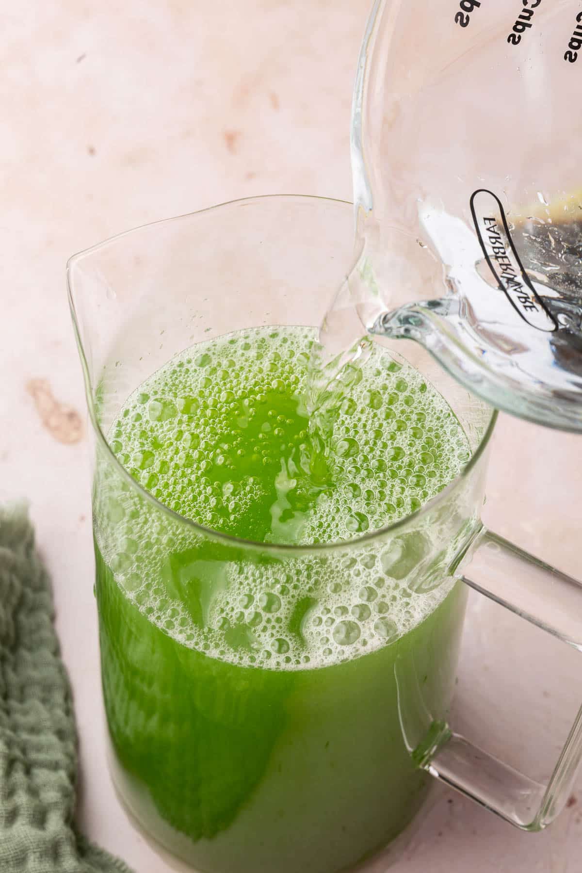 A measuring cup of water being poured into a pitcher of green cucumber lemonade.