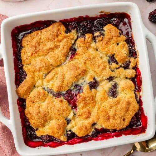 A square baking dish of blackberry cobbler with a bowl of blackberries peaking in.