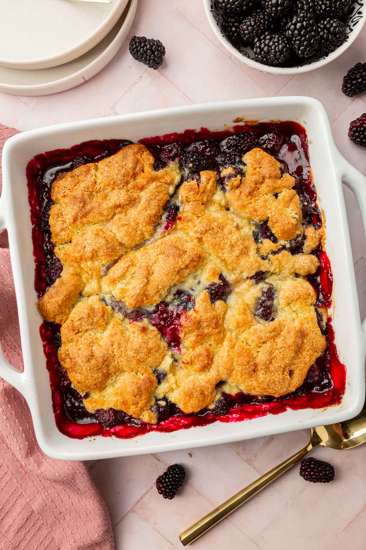 A square baking dish of blackberry cobbler with a bowl of blackberries peaking in.