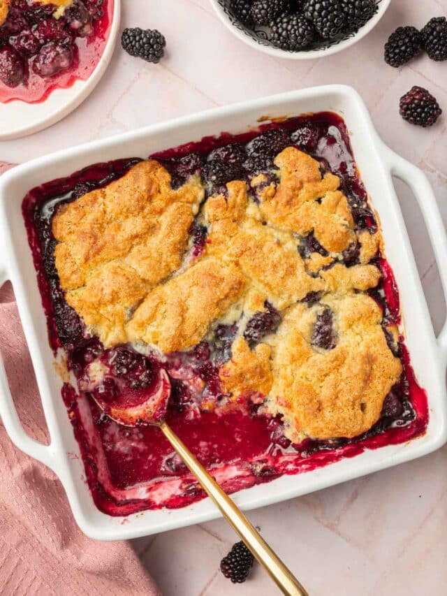 A square baking dish of gluten free blackberry cobbler with a spoon in it and a single serving on a plate peaking in and a small bowl of blackberries peaking in.