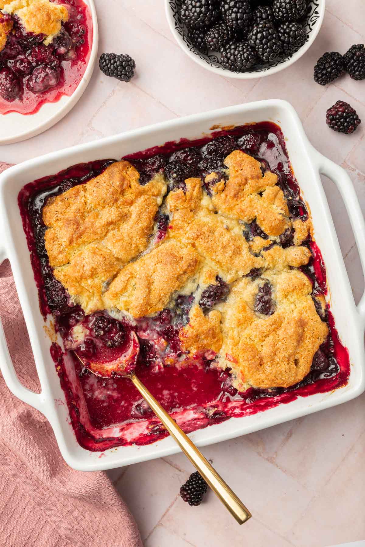 A square baking dish of gluten free blackberry cobbler with a spoon in it and a single serving on a plate peaking in and a small bowl of blackberries peaking in.