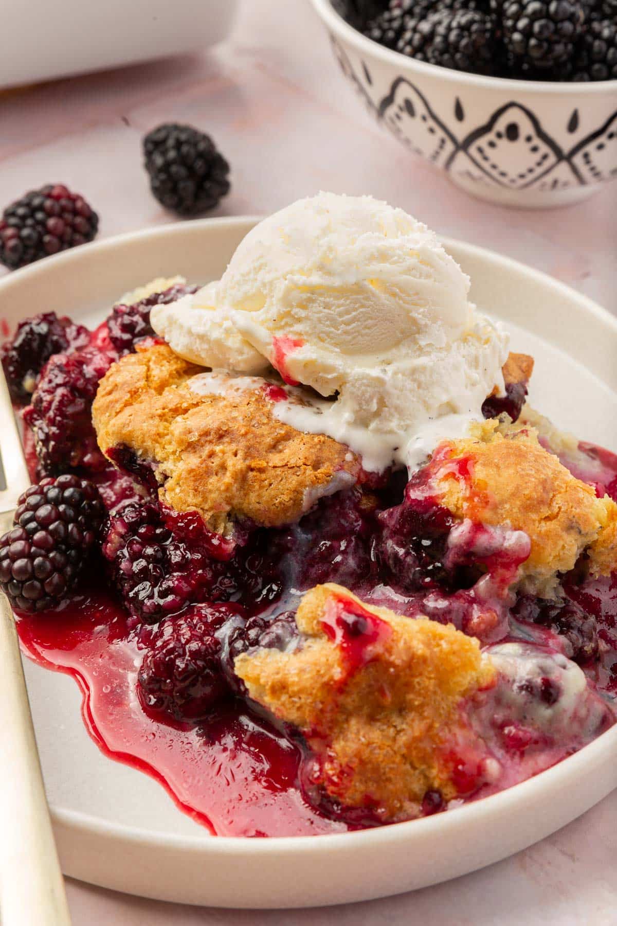 A single serving of gluten-free blackberry cobbler on a plate with a scoop of vanilla ice cream on top and a bowl of blackberries in the background.