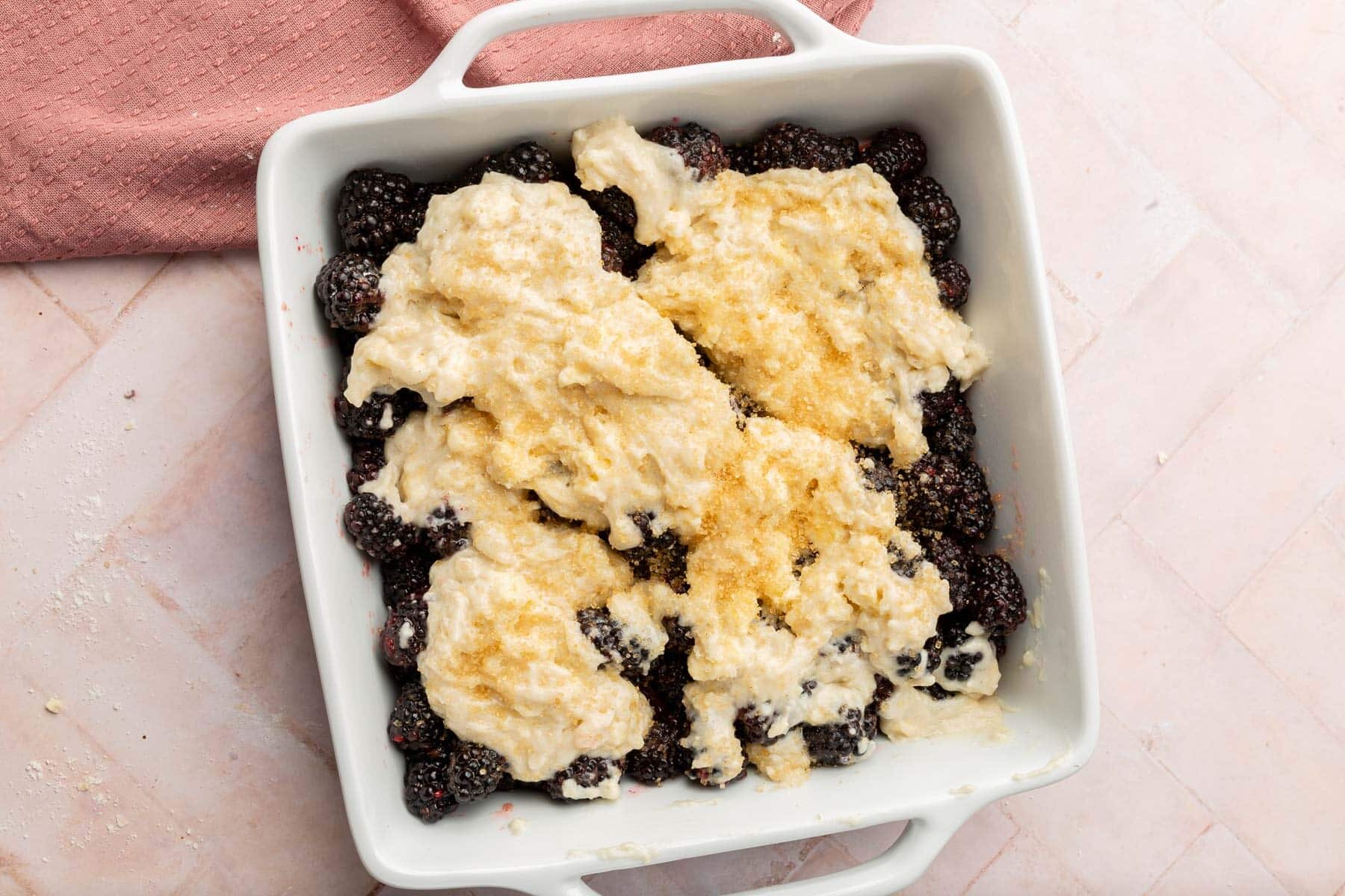 An overhead view of a square baking dish with blackberries topped with gluten-free cobbler batter and turbinado sugar before baking in the oven.