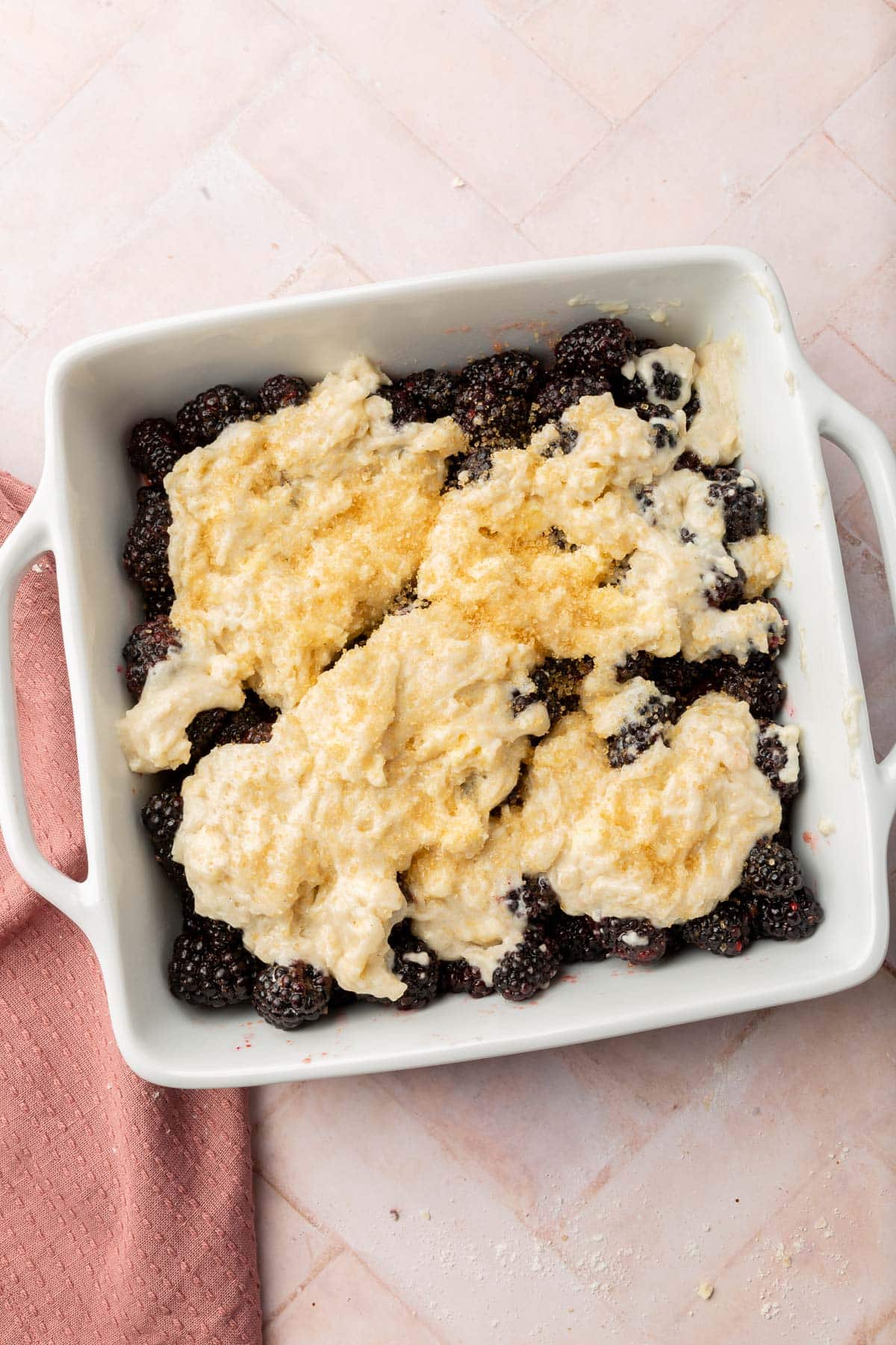 An overhead view of a square baking dish with blackberries topped with gluten-free cobbler batter and turbinado sugar before baking in the oven.