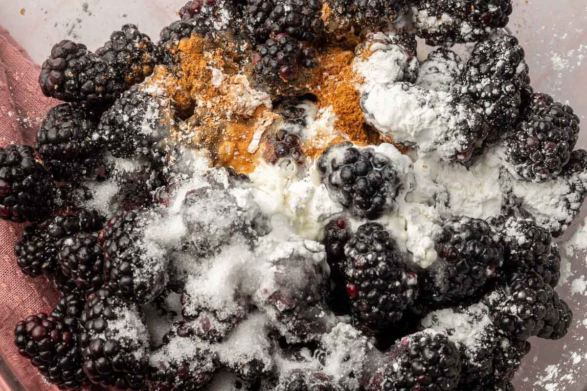 A glass mixing bowl with blackberries topped with granulated sugar, cornstarch, cinnamon and lemon juice before mixing together.