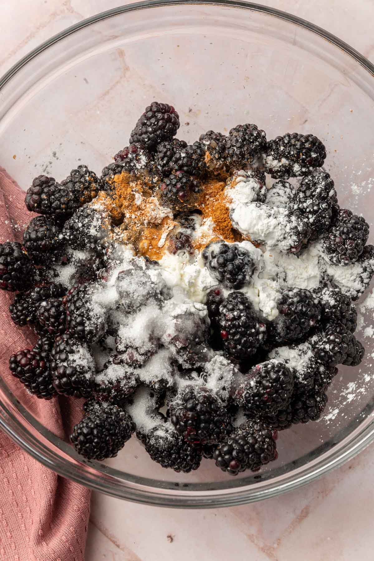 A glass mixing bowl with blackberries topped with granulated sugar, cornstarch, cinnamon and lemon juice before mixing together.