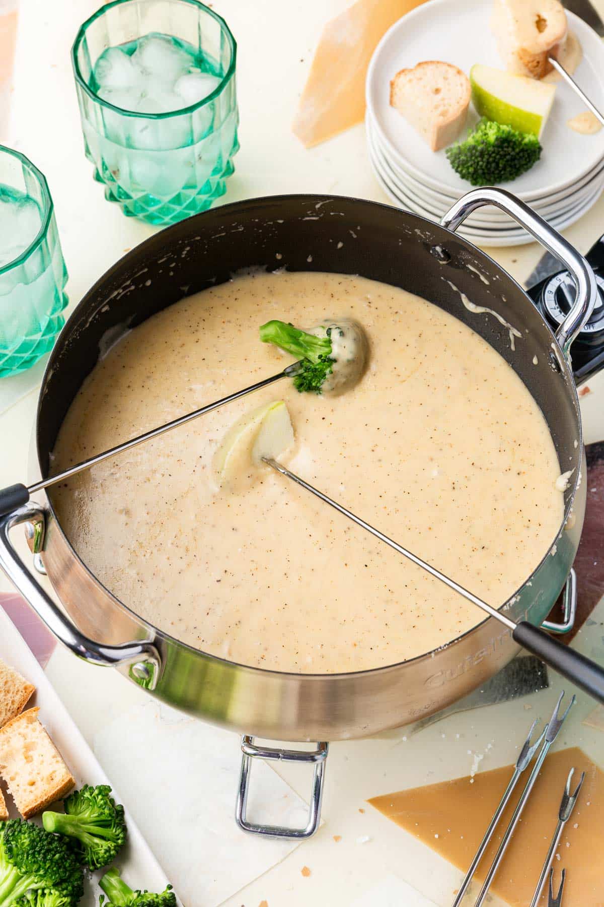 An electric fondue pot of glute-free cheese fondue that has a skewer of broccoli and a skewer of apple dipped into it with additional plates of dippers to the side.