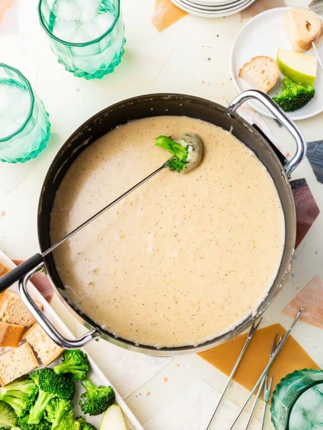 An electric fondue pot of gluten-free cheese fondue with a skewered piece of broccoli dipped into it with a platter of gluten-free bread, broccoli and apples to the side.