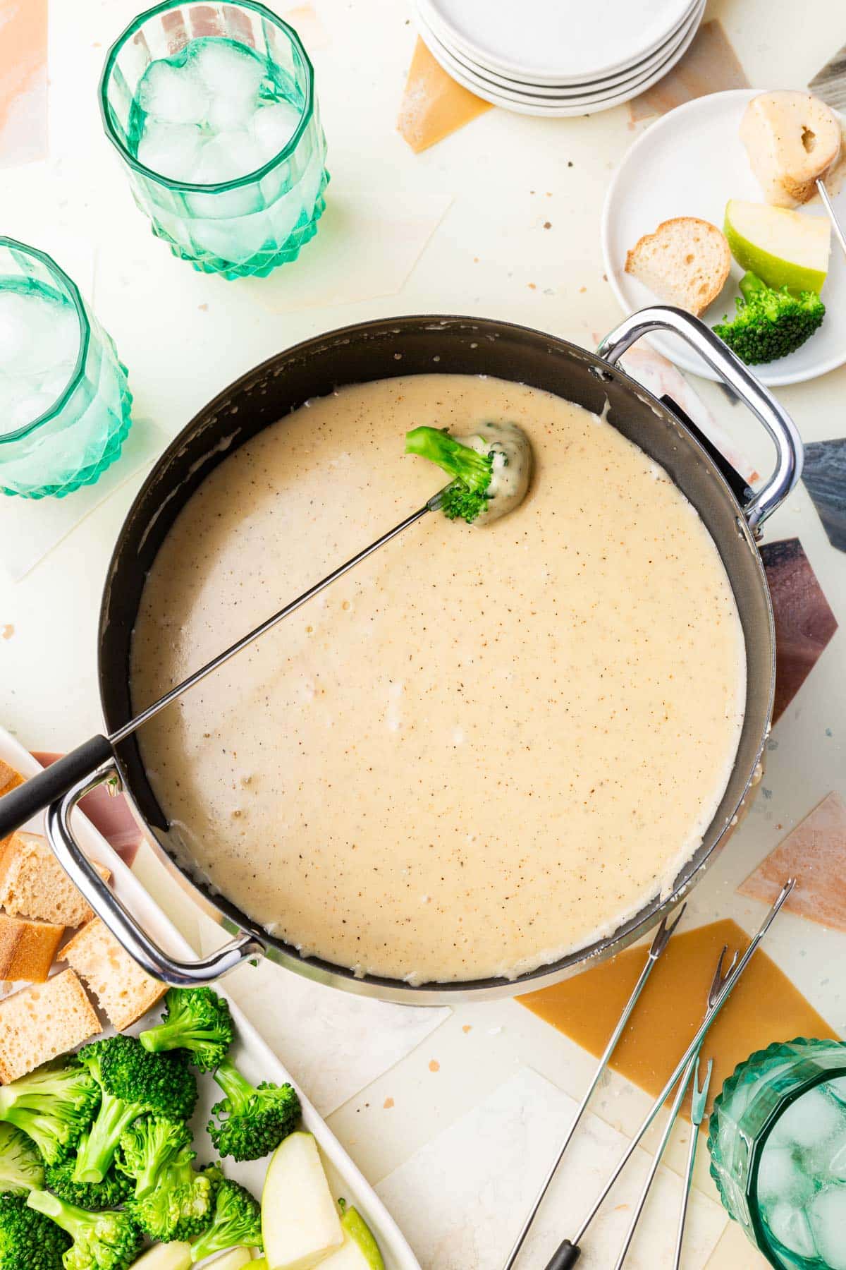 An electric fondue pot of gluten-free cheese fondue with a skewered piece of broccoli dipped into it with a platter of gluten-free bread, broccoli and apples to the side.