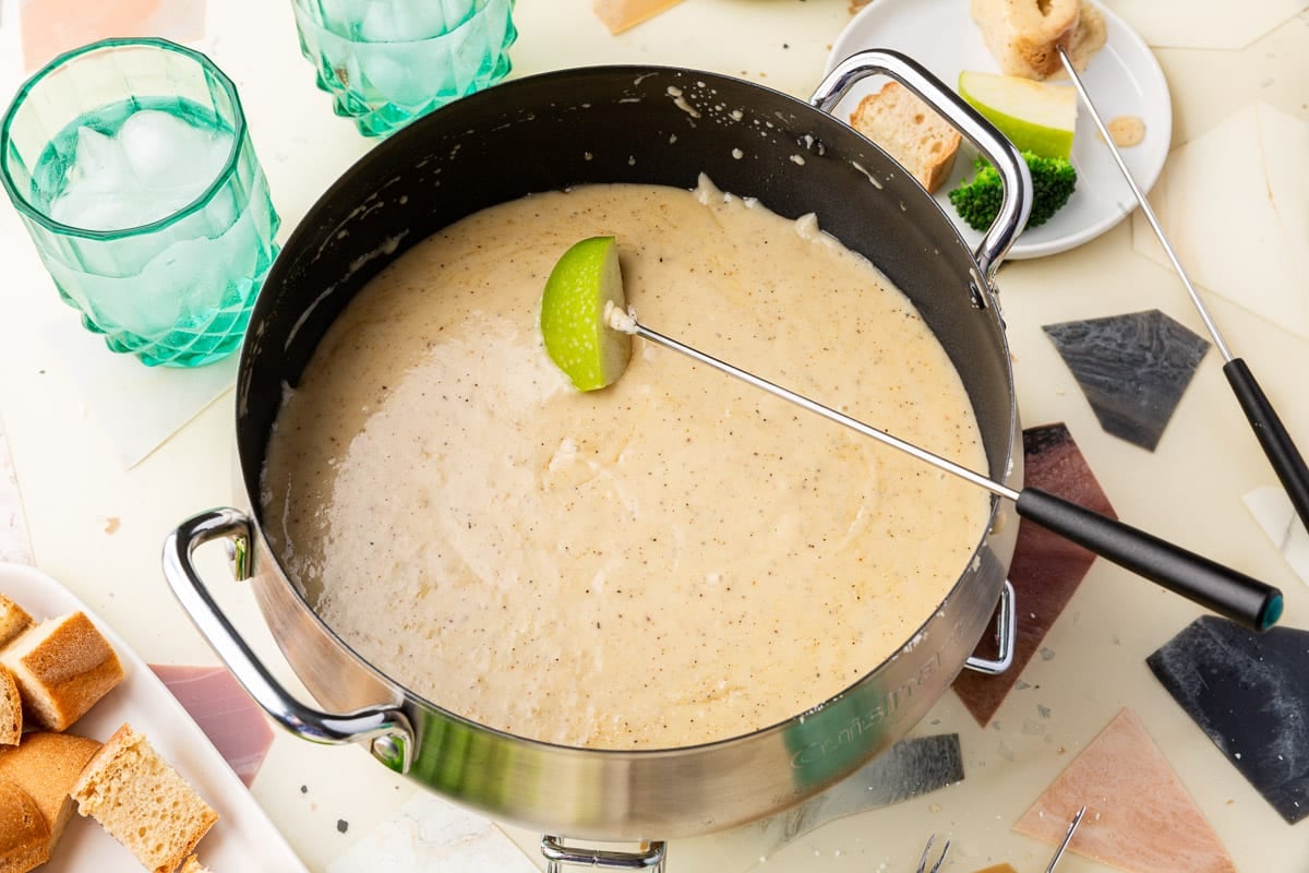 A fondue fork skewered with green apple that is dipped into an electric fondue pot of cheese fondue with plates of bread, broccoli and apple to the side.