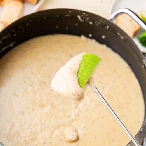 A fondue fork skewered with a piece of granny smith apple that has cheese fondue dripping off it into an electric fondue pot.