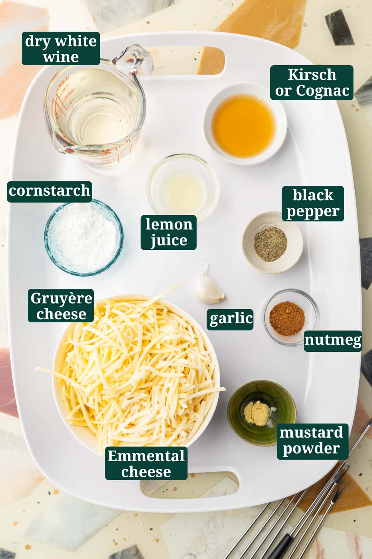 Ingredients in small bowls to make gluten-free cheese fondue including dry white wine, cornstarch, lemon juice, Kirsh, black pepper, Gruyère cheese, Emmental cheese, a garlic clove, nutmeg, and mustard powder with text overlays over each ingredient to say what it is.