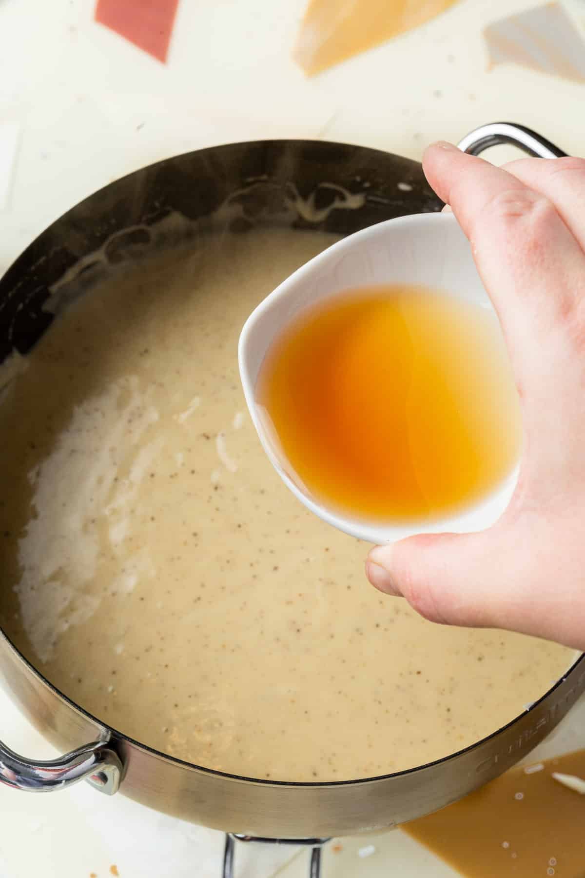 A hand pouring a ramekin of Cognac into a fondue pot of melted cheese.