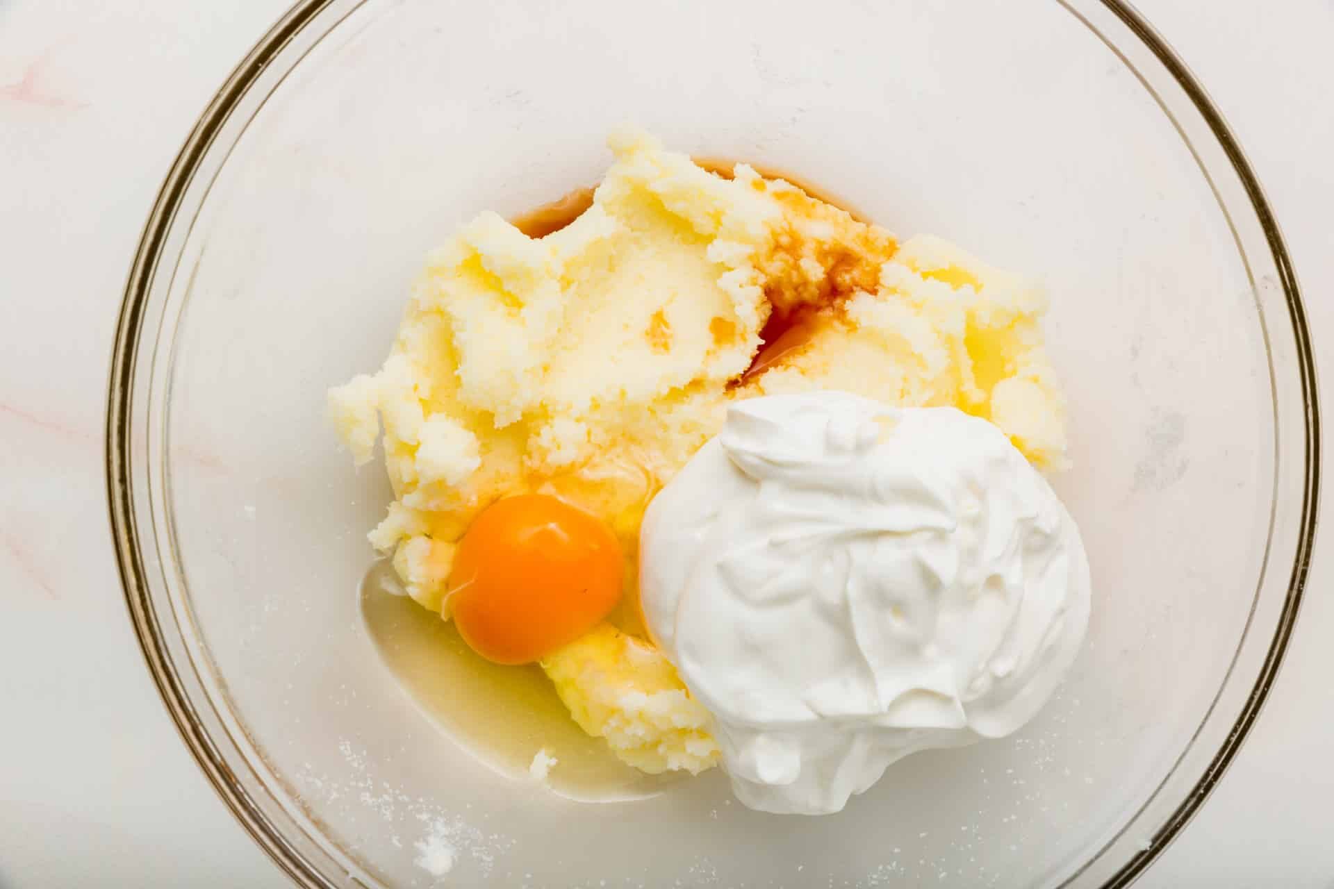 A glass mixing bowl with sour cream, eggs, vanilla, and a creamed sugar and butter mixture.
