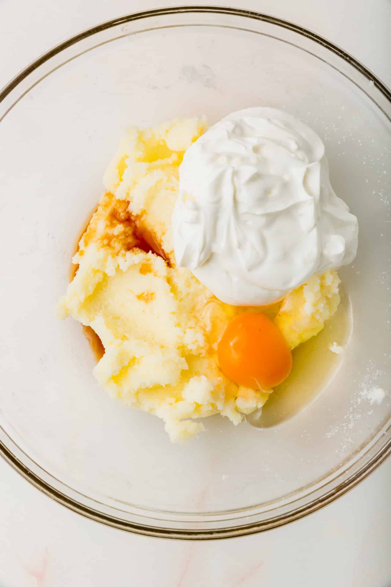A glass mixing bowl with sour cream, eggs, vanilla, and a creamed sugar and butter mixture.