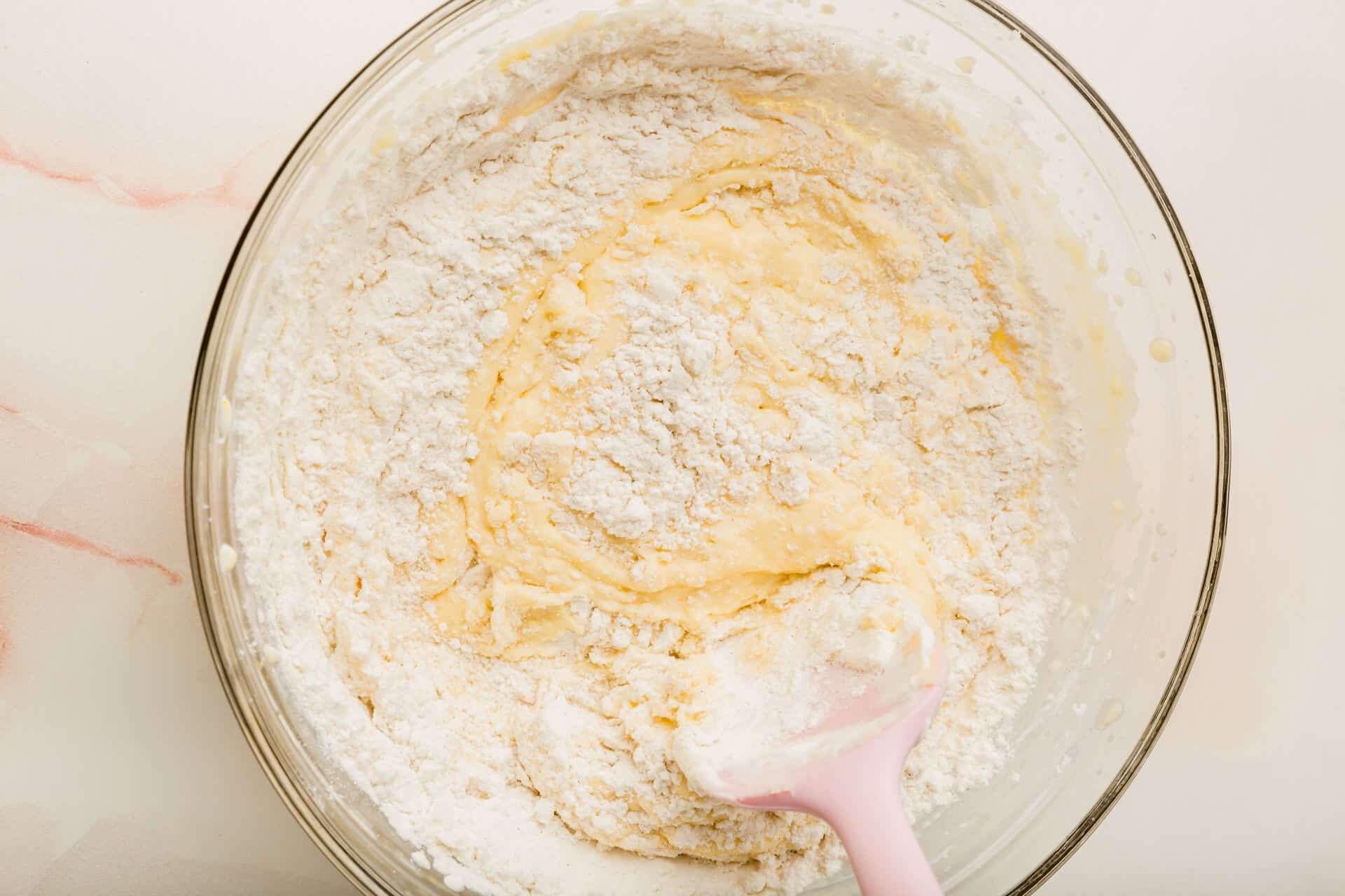 A glass mixing bowl of a gluten-free flour blend being mixed together with a wet batter with a pink spatula.