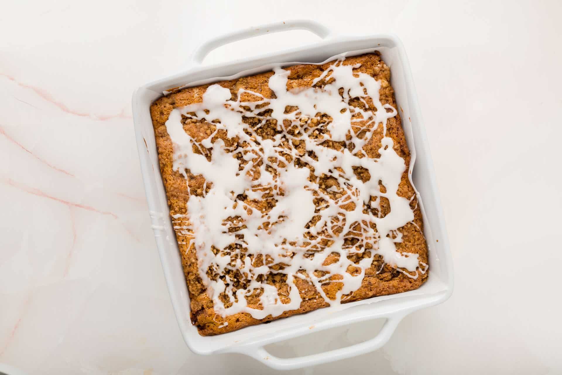 An overhead view of a gluten-free coffee cake in a white square baking dish with a drizzled glaze.