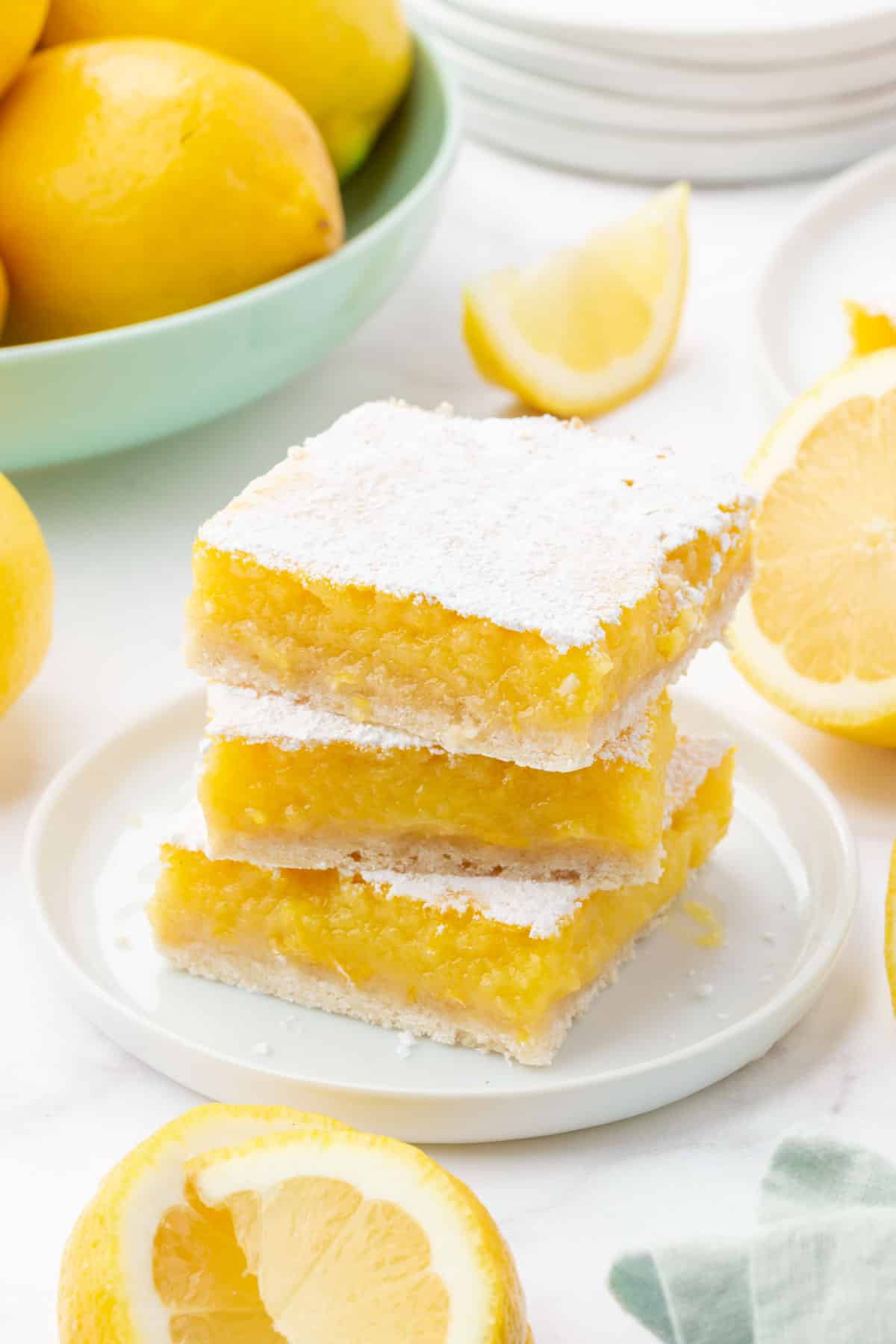 A stack of three gf lemon bars on a dessert plate with a bowl of lemons in teh background and lemon slices around the lemons.