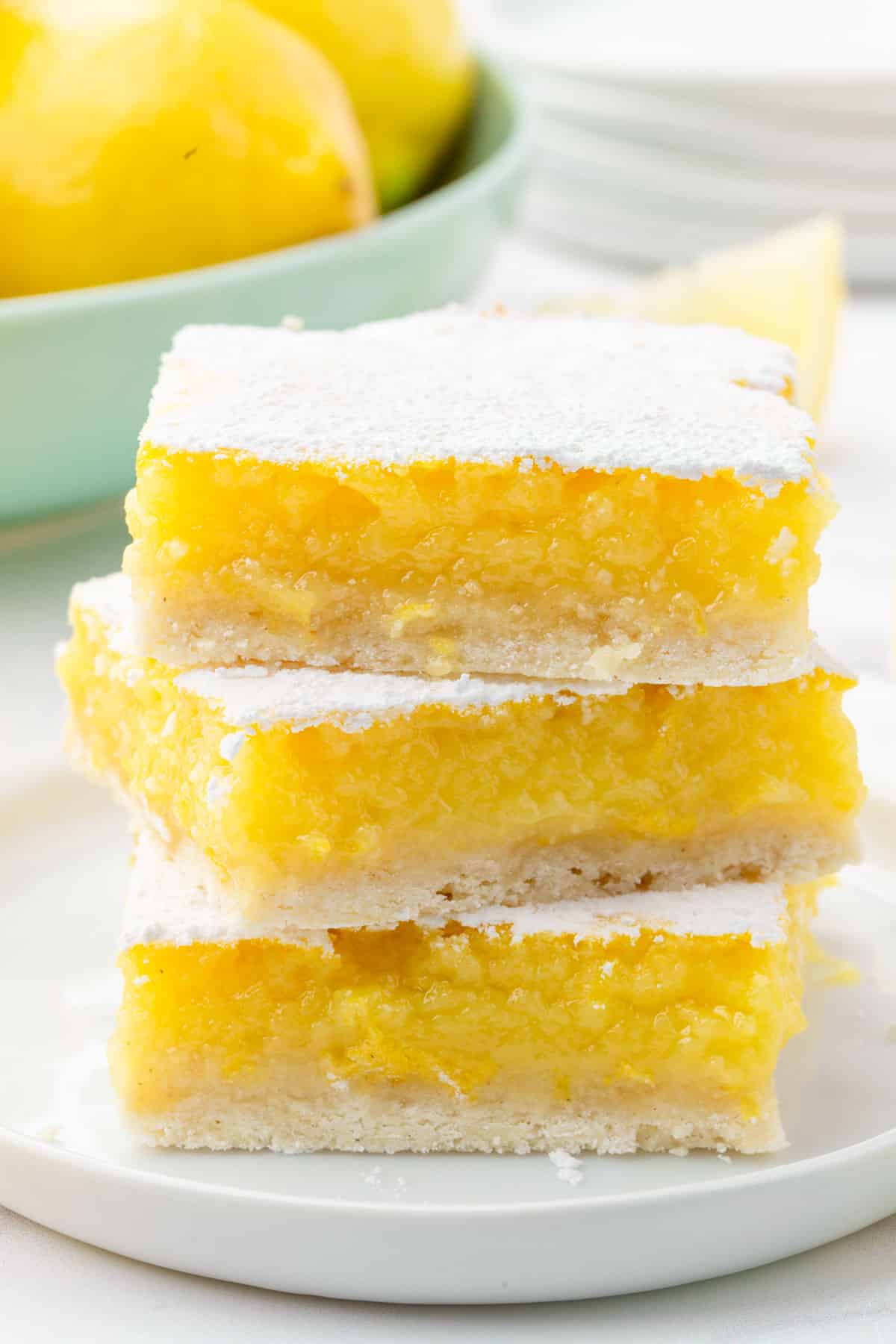 A stack of three gluten free lemon bars on a dessert plate with a bowl of lemons in the background.