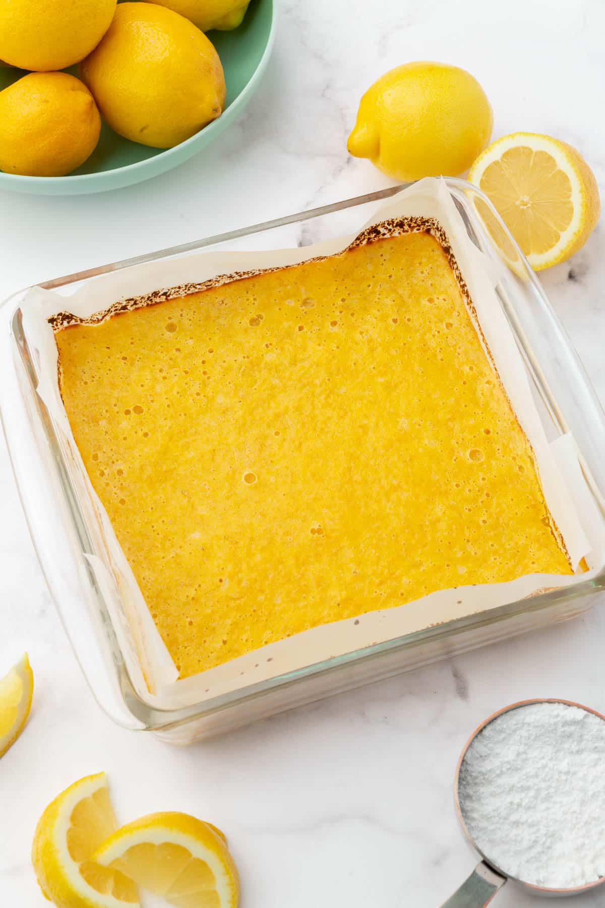 A square glass baking with baked gluten free lemon bars in it with a bowl of lemons and cut lemons in the background.