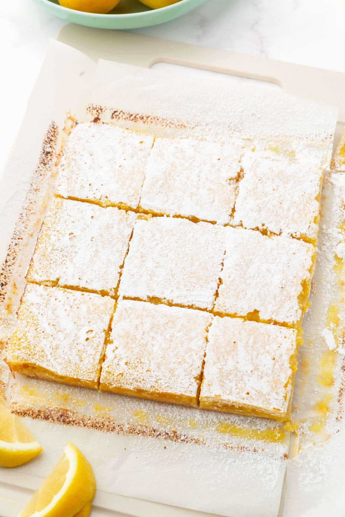 A large slab of gluten free lemon squares cut into 9 equal squares on a cutting board and dusted with powdered sugar.