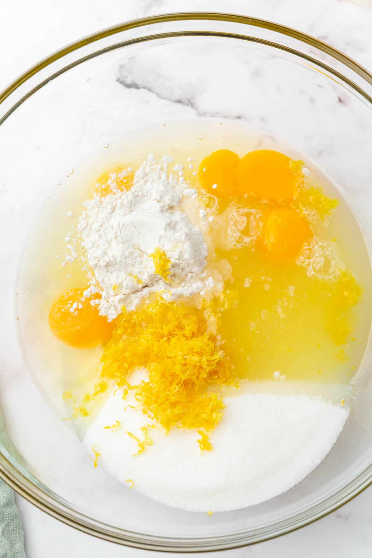 A glass mixing bowl with eggs, lemon juice, lemon zest, gluten-free flour, and granulated sugar before whisking together.