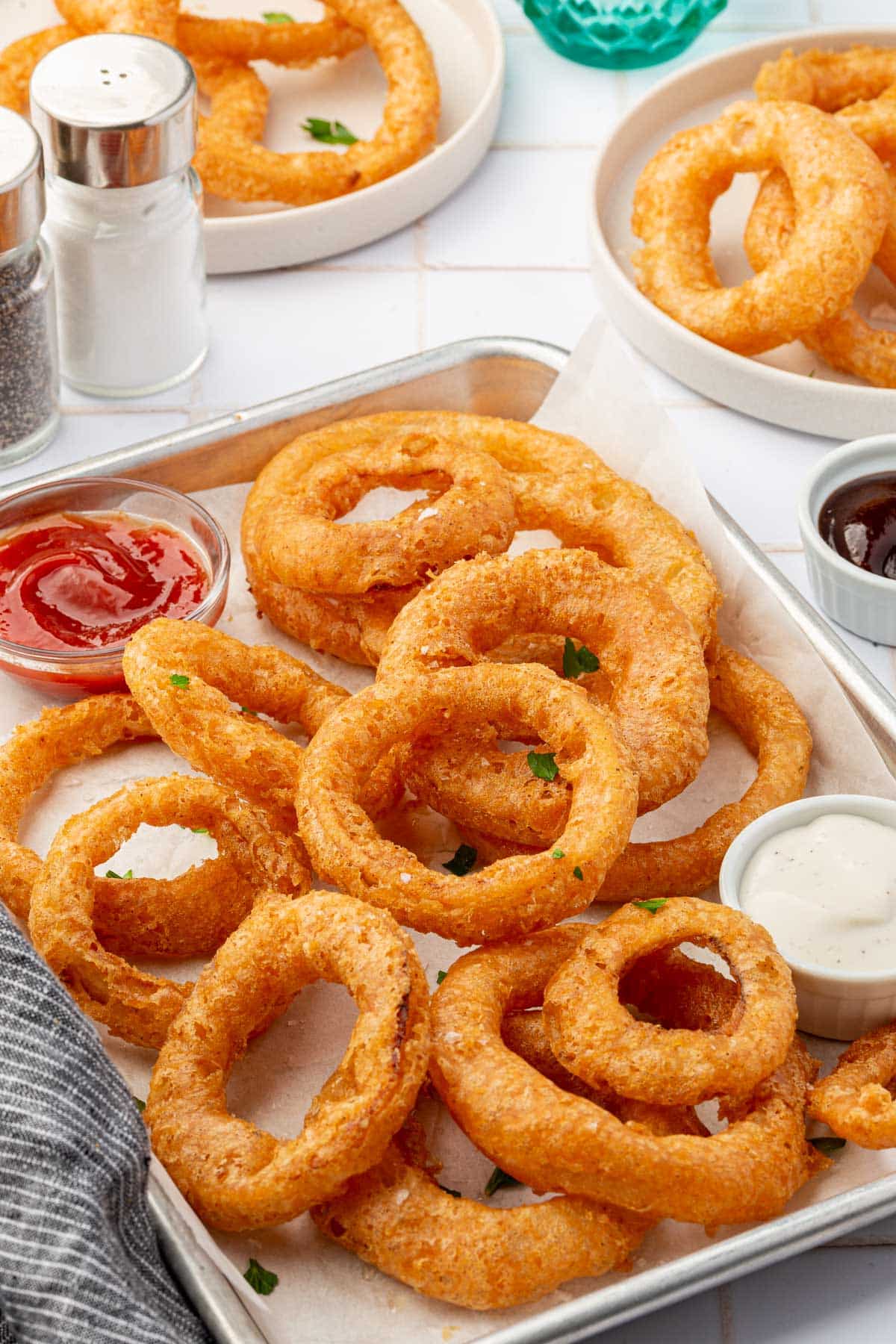 A parchment lined baking sheet of gluten free onion rings with ramekeins of ketchup and ranch with additional plates of onion rings in the background.