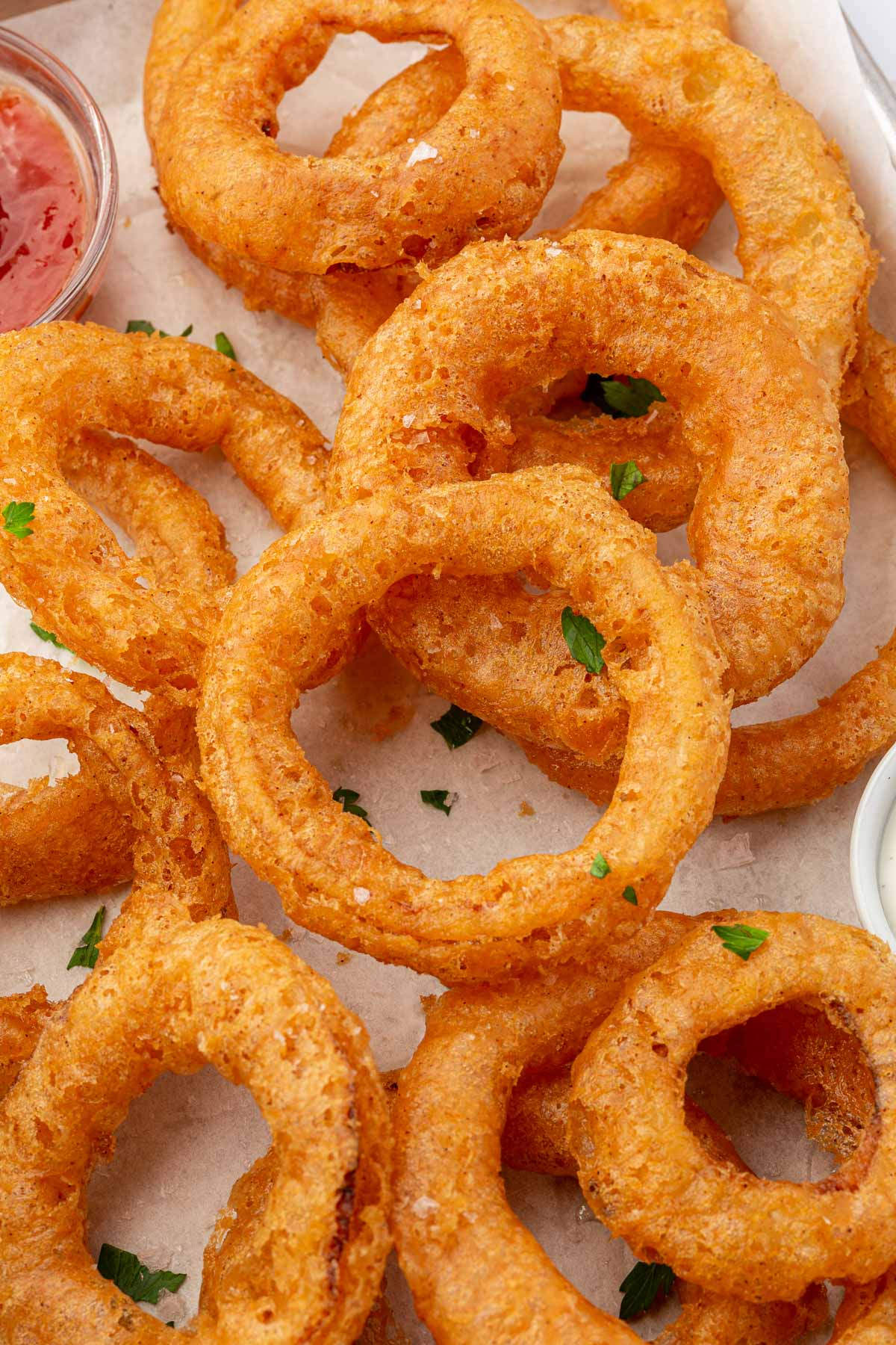 A close up of gluten free onion rings topped with parsley on a parchment lined baking sheet with ketchup peaking in.