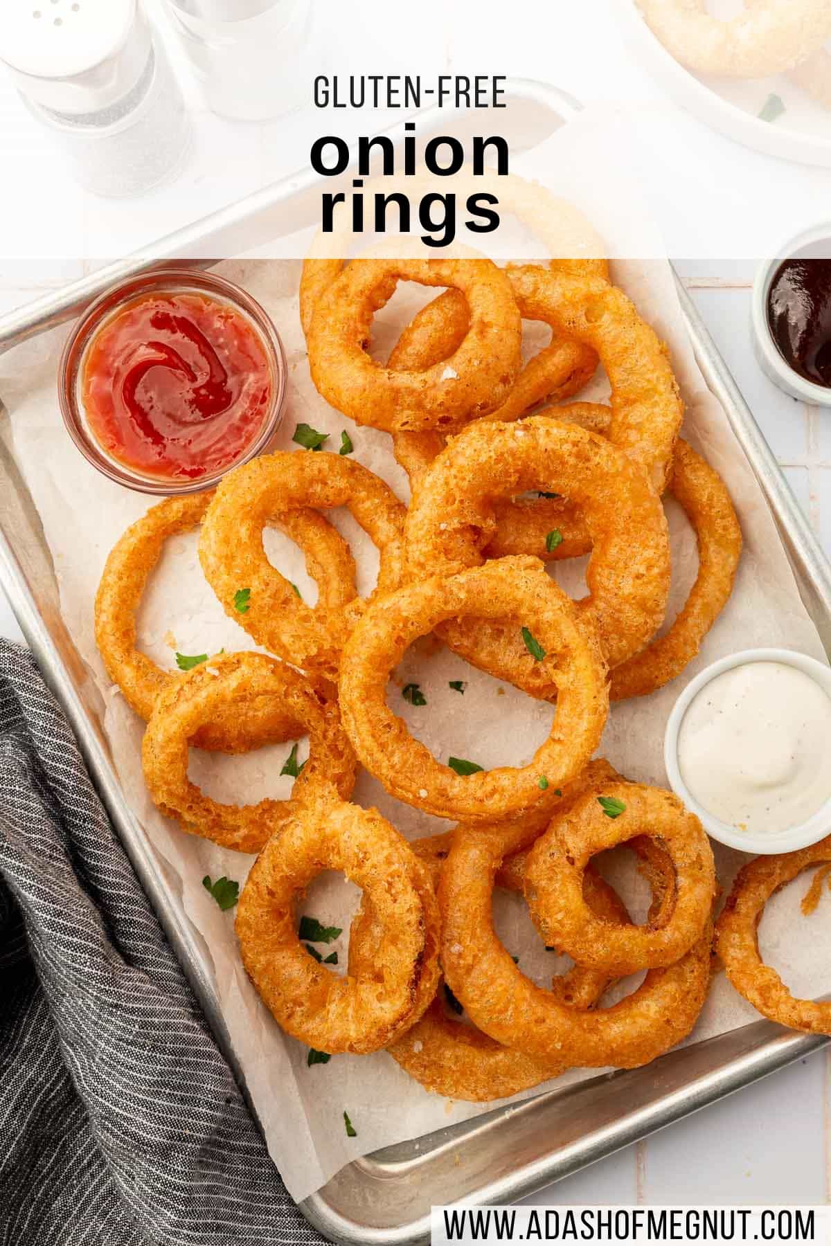 An overhead view of gf onion rings on a parchment lined baking sheet with bowls of ranch and ketchup.