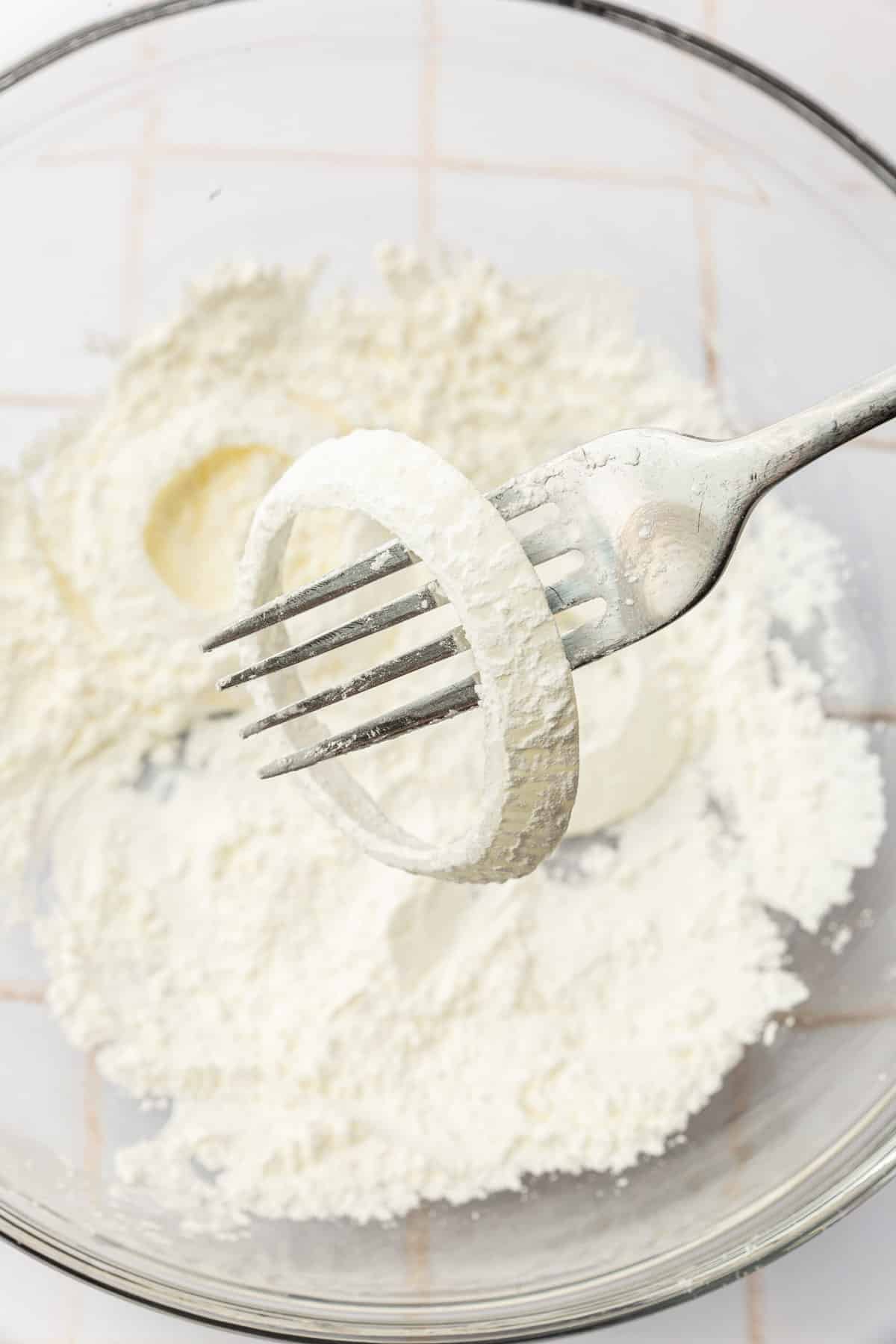 A cornstarch dipped onion ring on a fork over a bowl of additional cornstarch.