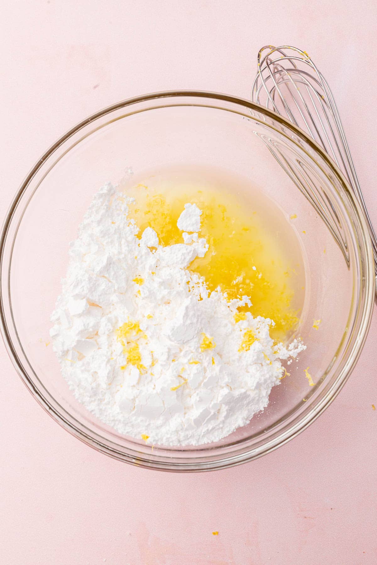 A glass mixing bowl with powdered sugar, lemon juice and lemon zest before mixing together.