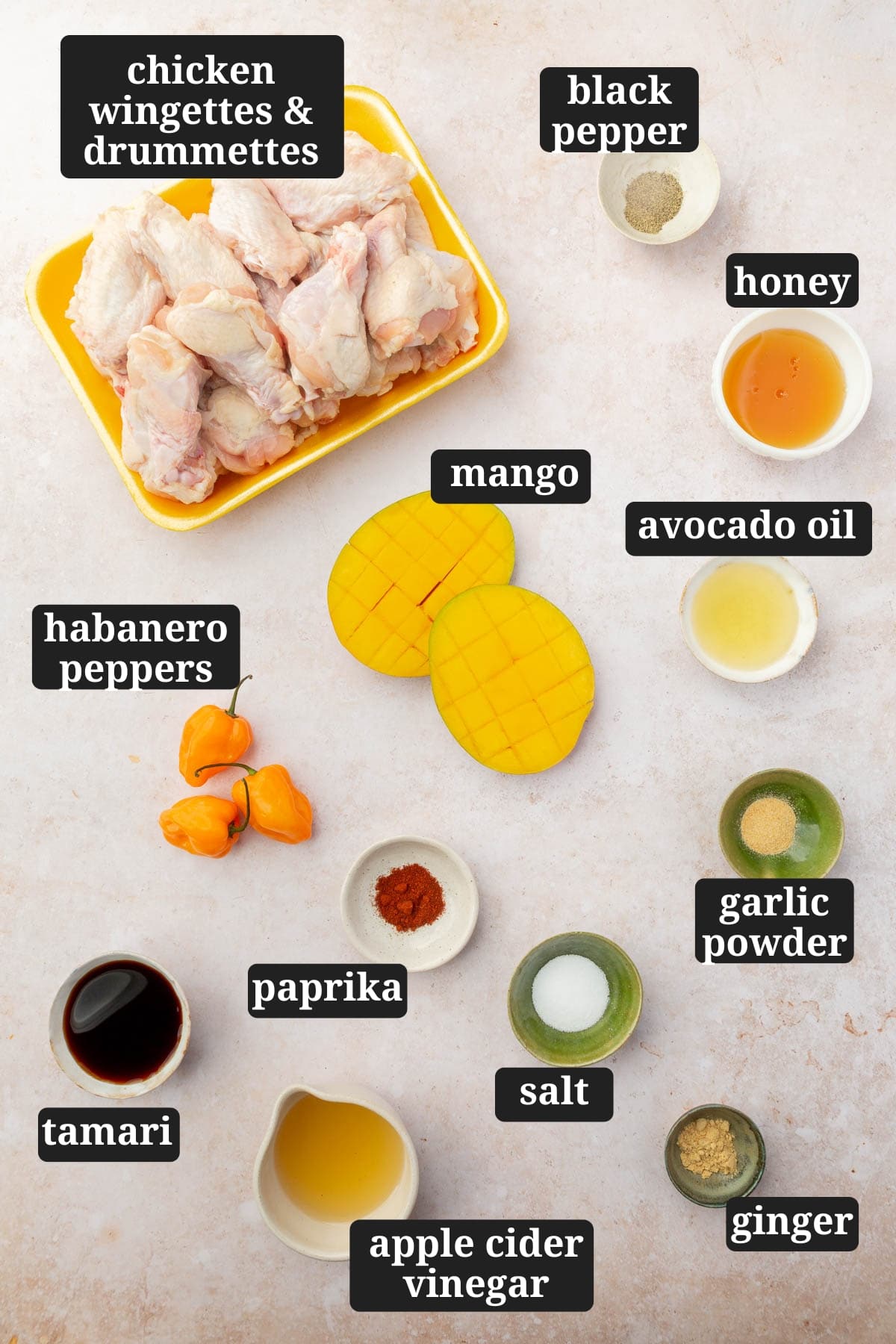 An overhead view of ingredients in small bowls to make mango habanero chicken wings, including chicken drumettes and flats, black pepper, honey, apple cider vinegar, mango, habanero peppers, paprika, salt, garlic powder, ginger, avocado oil, and tamari with text overlays over each ingredient.