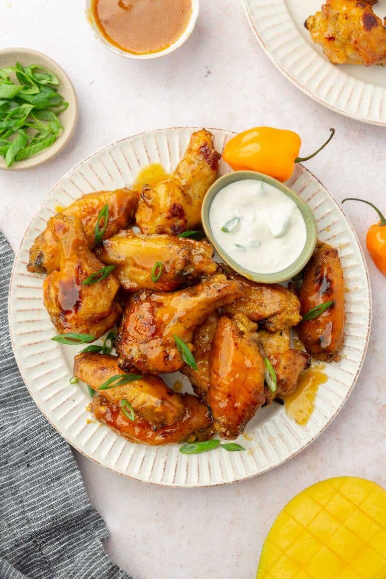 A plate of baked mango habanero wings topped with green onion with a ramekin of bleu cheese dressing on the side and habanero peppers on the plate.