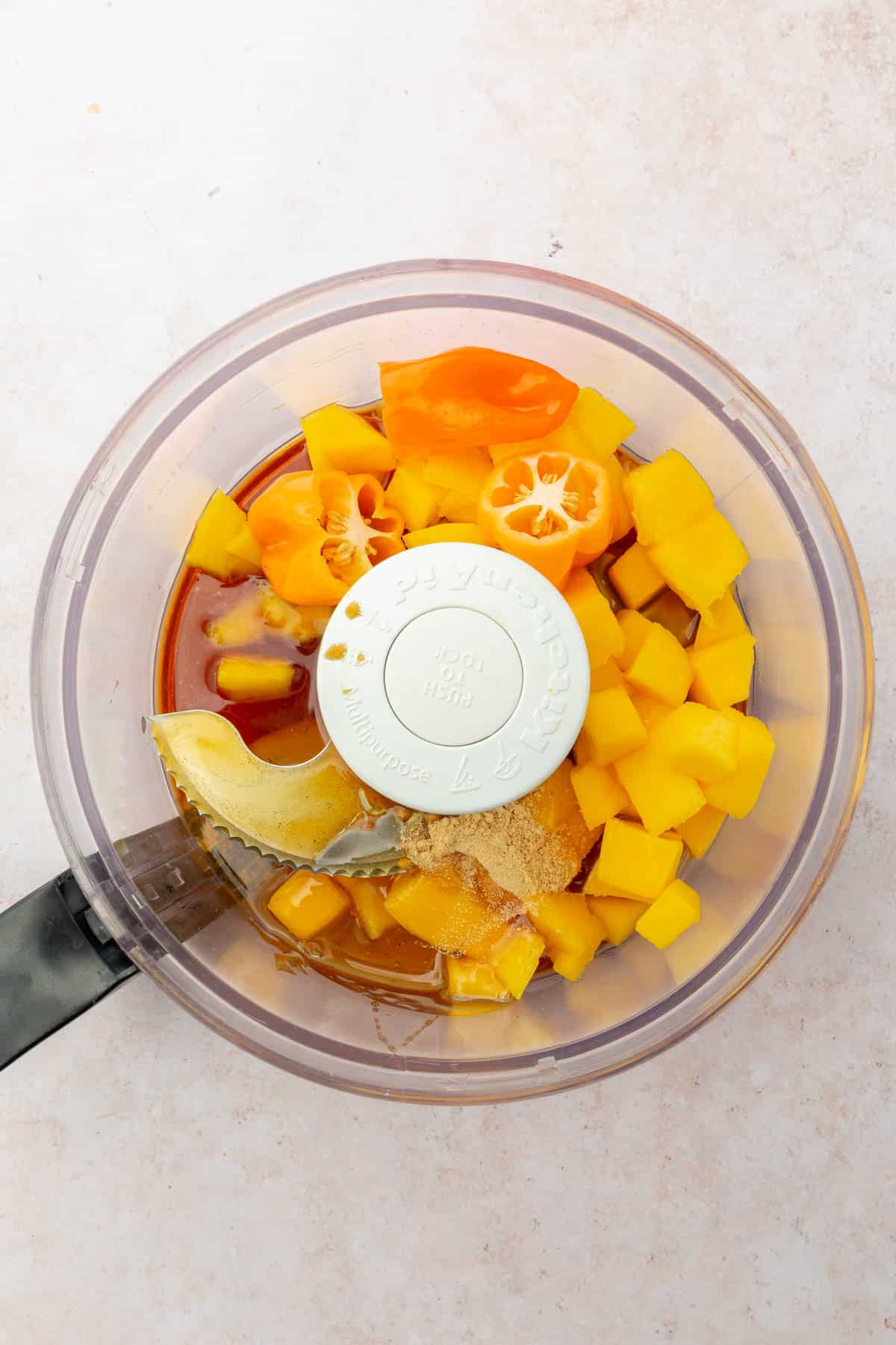 A food processor with habanero peppers, mango cubes, honey, ginger powder, tamari sauce, and apple cider vinegar.