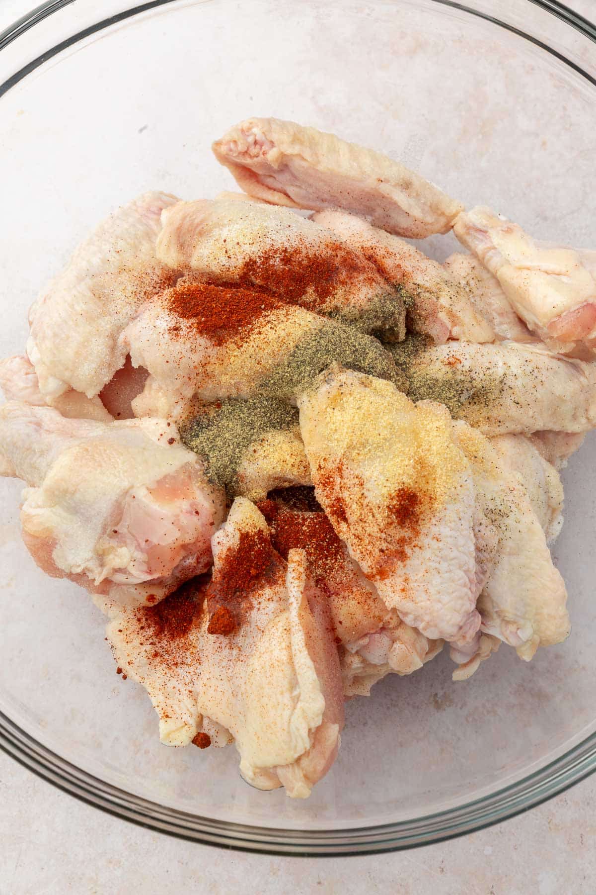 A glass mixing bowl with raw chicken wings topped with paprika, garlic powder, salt, and pepper before mixing together.