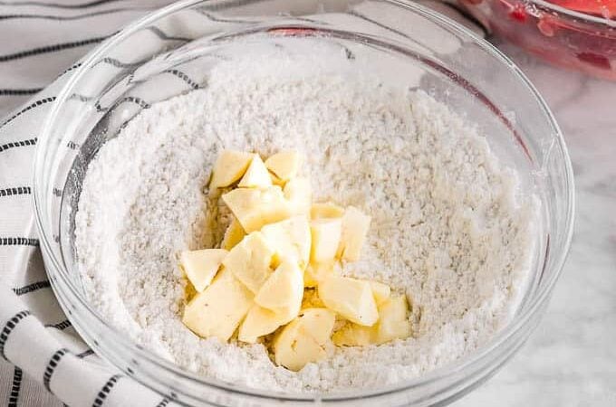 A clear glass bowl containing flour and chunks of butter, placed on a striped kitchen towel with part of another bowl visible in the background.