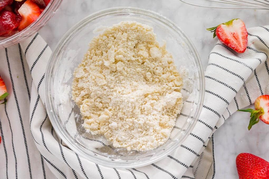 A glass bowl filled with crumbly streusel topping sits on a striped kitchen towel, with fresh sliced strawberries scattered nearby on a marble surface.