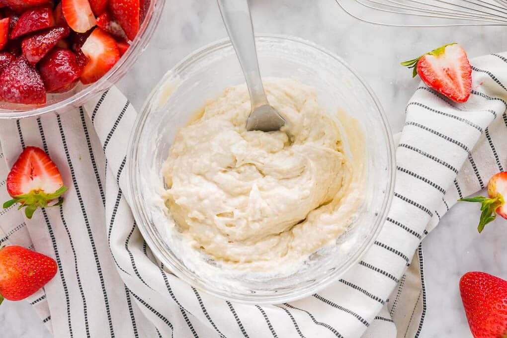 A glass bowl filled with a creamy batter and a spoon sits on a striped cloth. Fresh strawberries, some halved, are scattered around, with a bowl of chopped strawberries nearby.