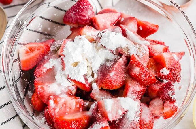A glass bowl filled with chopped strawberries mixed with sugar and flour sits on a striped cloth, surrounded by fresh strawberries, a lemon half, and a wooden measuring cup.