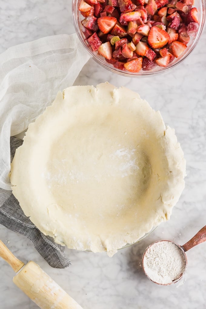 Gluten-free pie dough that has been pressed into a pie plate with a bowl of strawberry rhubarb filling to the side.