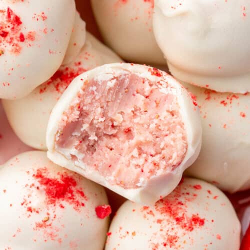 A close up shot of a strawberry truffle with a bite taken out of it sitting on top of a plate of more white chocolate strawberry truffles.