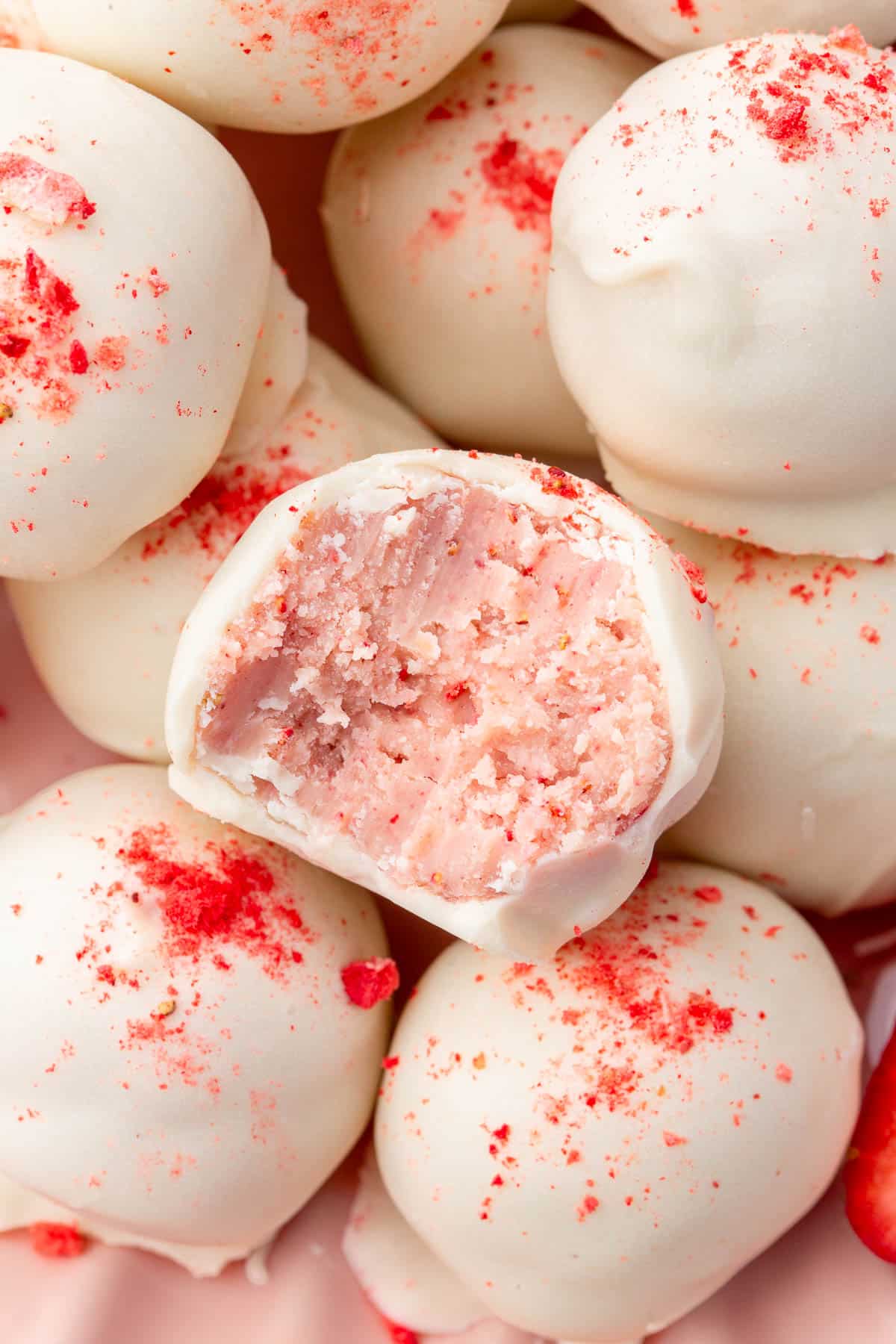 A close up shot of a strawberry truffle with a bite taken out of it sitting on top of a plate of more white chocolate strawberry truffles.