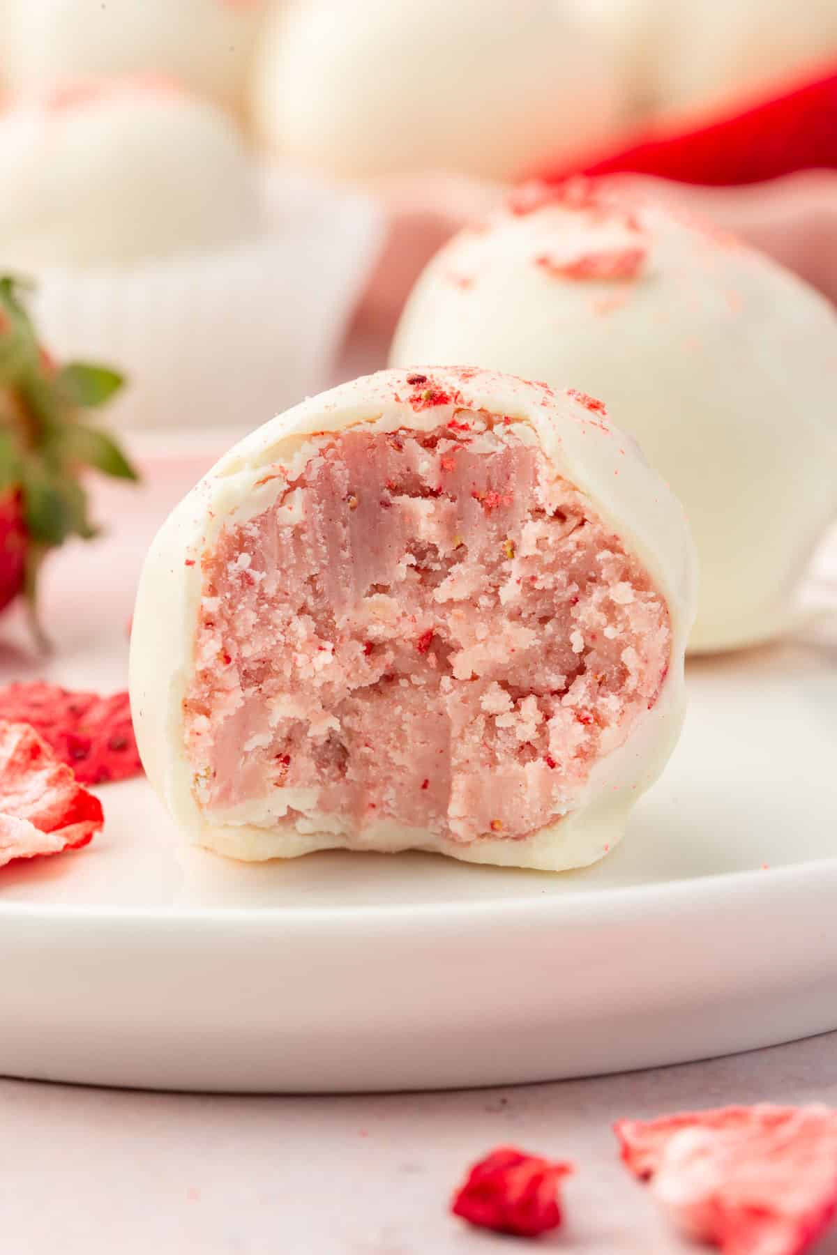 A single strawberry truffle coated in white chocolate on a plate with additional truffles in the background.