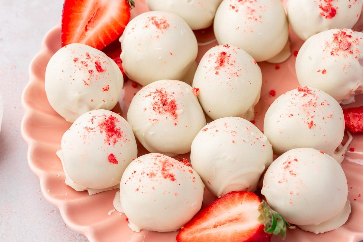 A pink scalloped plate with white chocolate strawberry truffles and a few fresh strawberry halves on it.