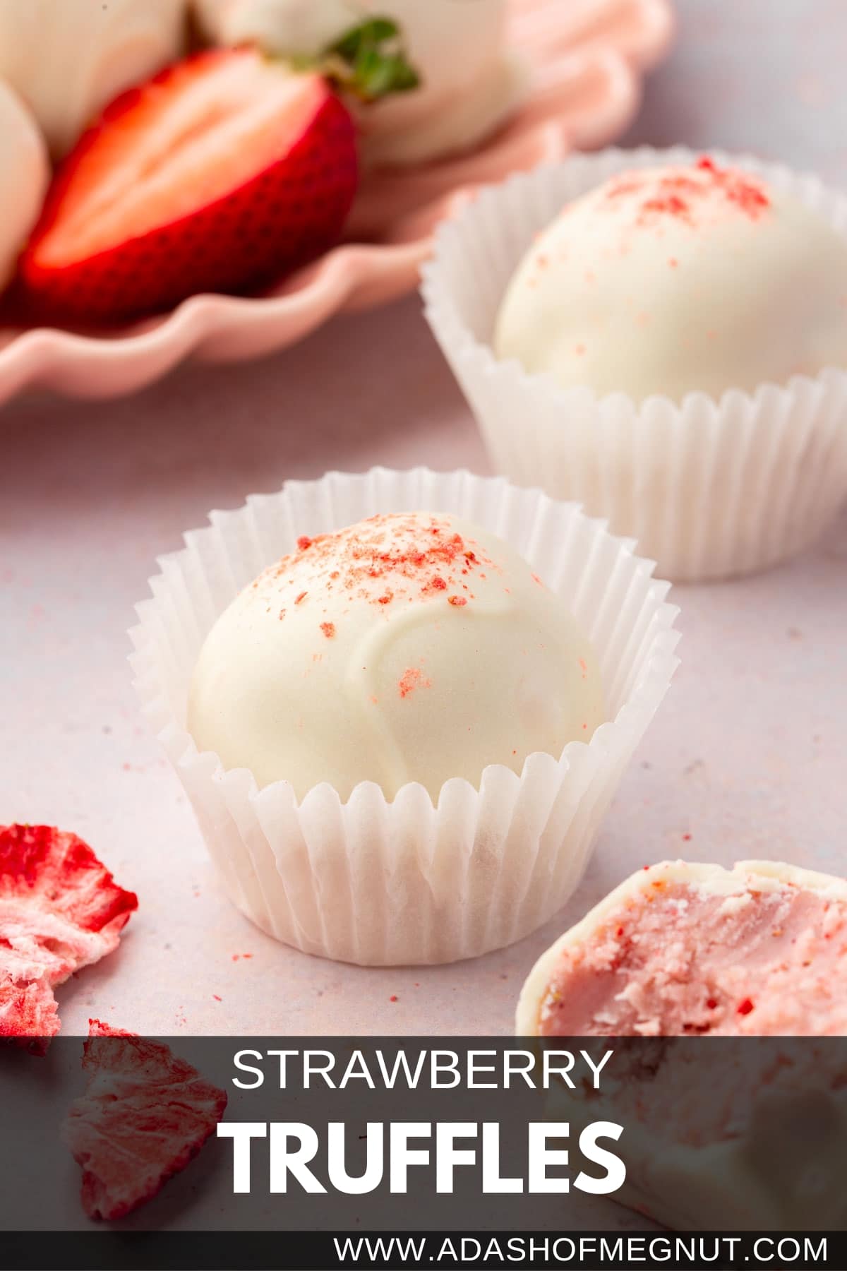 Two white chocolate strawberry truffles in paper cups with a plate of additional truffles and fresh strawberries in the background.