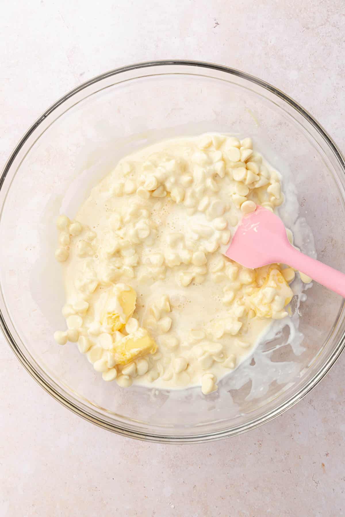 A glass mixing bowl with white chocolate chips, butter and heavy cream with a pink spatula.