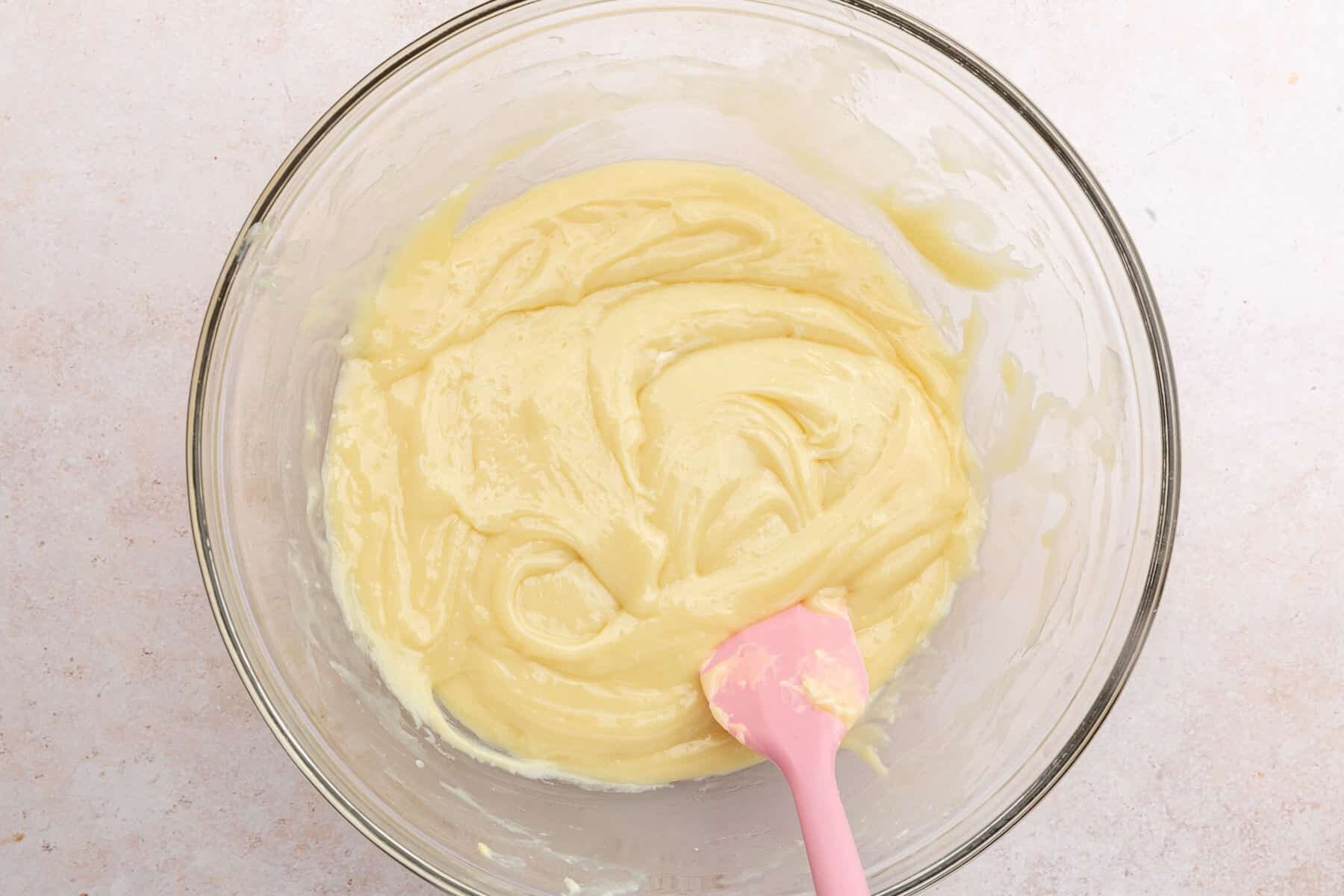 A glass mixing bowl with a melted white chocolate mixture and a pink spatula.