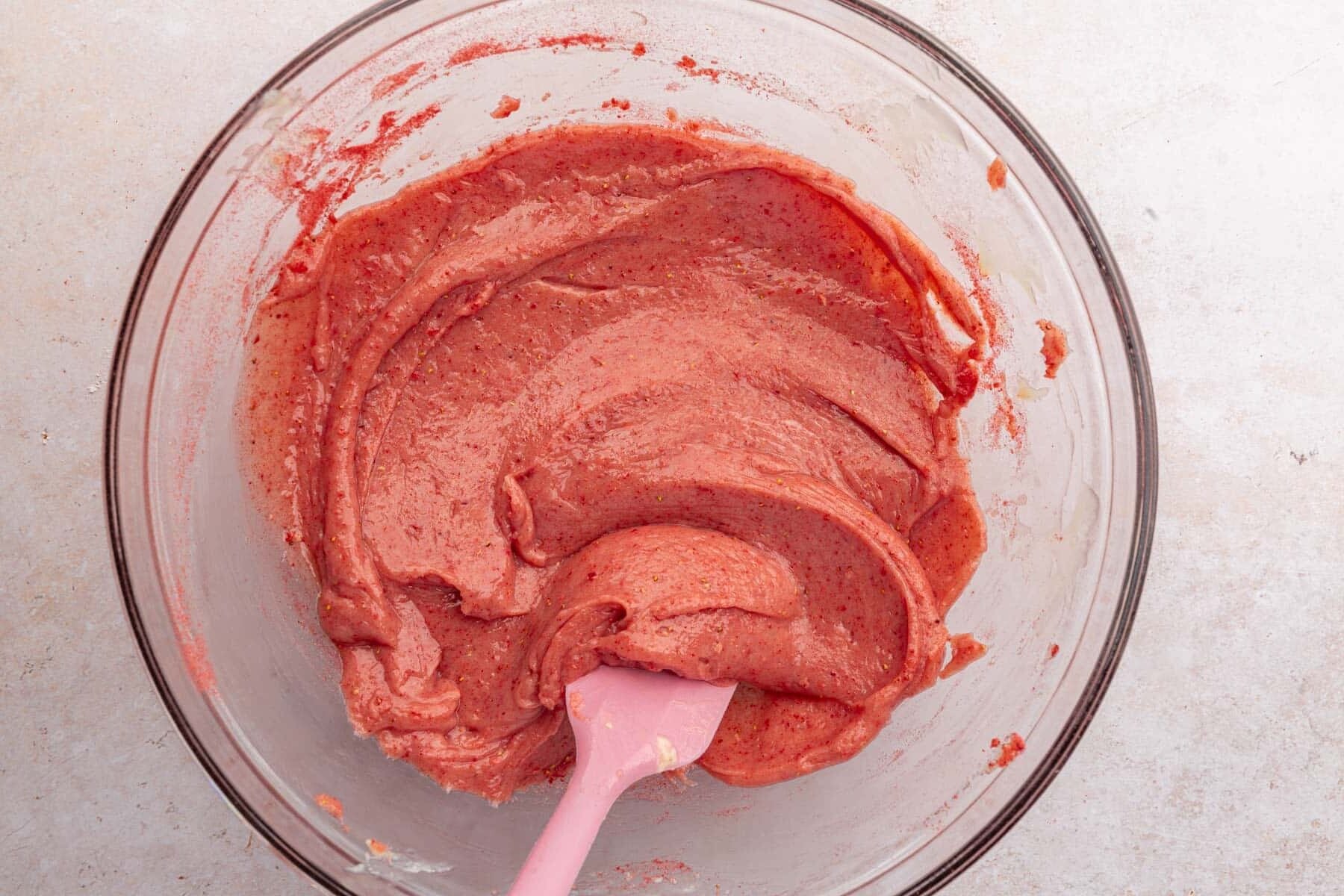 A glass mixing bowl with a strawberry dough mixture and a pink spatula.