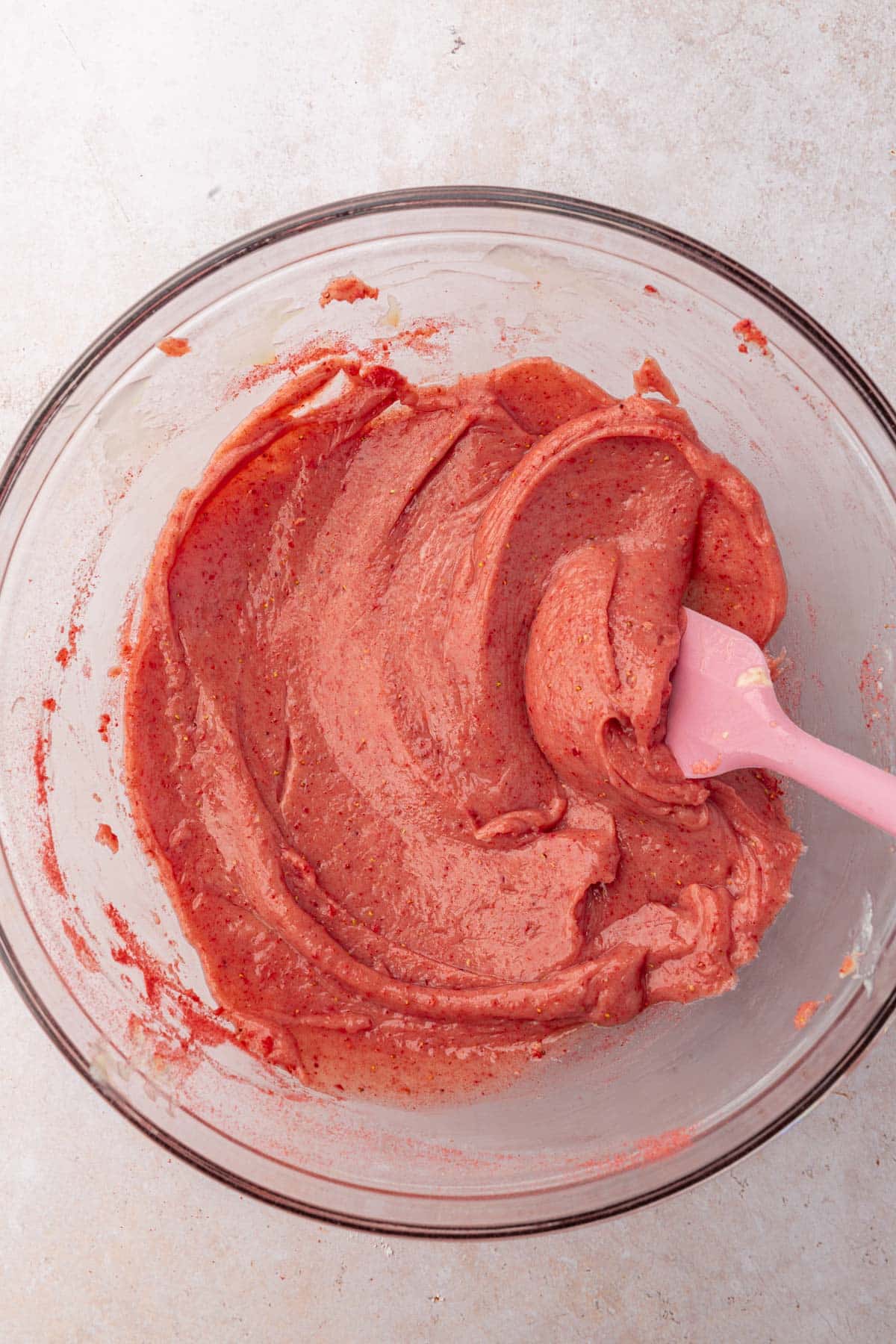 A glass mixing bowl with a strawberry dough mixture and a pink spatula.